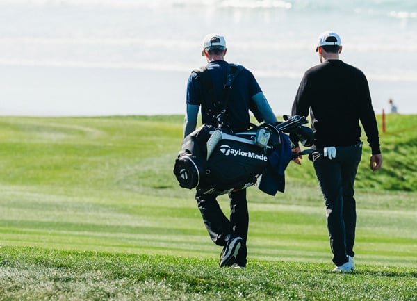 A golfer and his caddie (carrying a TaylorMade golf bag) walk away from the camera on a green fairway toward the bright ocean and a white sand beach.