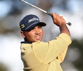 A close-up of golfer Jason Day mid-swing, wearing a yellow knit sweater and a blue