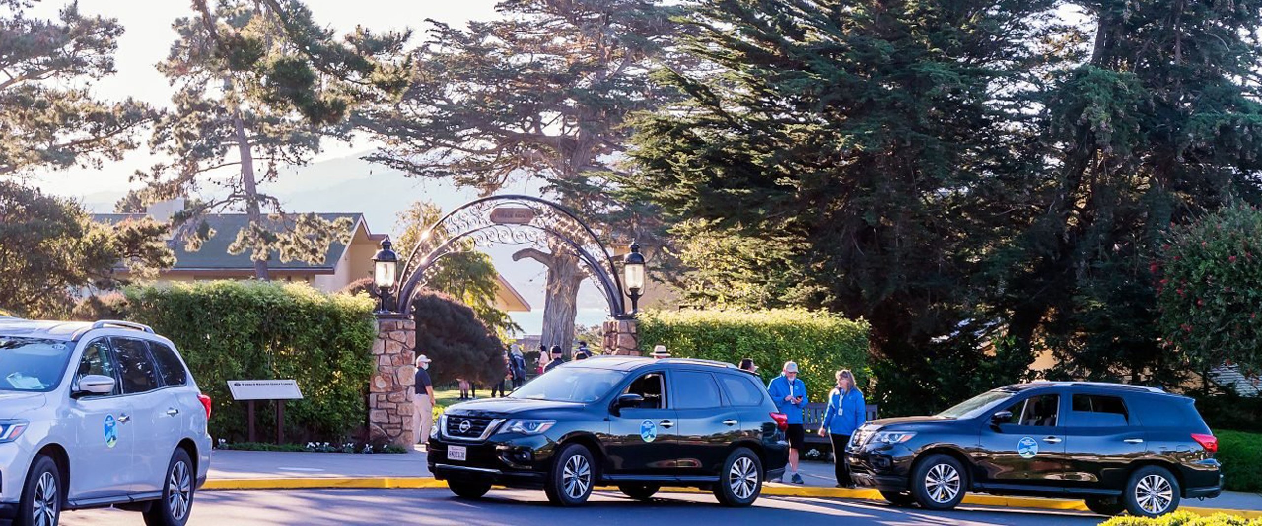 The entrance to a parking area for the Pro-Am, showing several dark SUVs driving through a wrought-iron archway with greenery and mature trees surrounding the road.