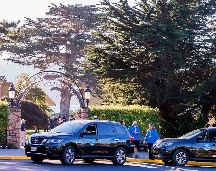 The entrance to a parking area for the Pro-Am, showing two dark SUVs driving through a decorative wrought-iron archway surrounded by large trees and volunteers in blue jackets.