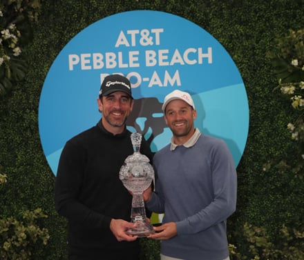 The winning Pro-Am team of Aaron Rodgers and Ben Silverman holding the crystal trophy in front of a blue and white AT&T Pebble Beach Pro-Am logo backdrop surrounded by green foliage.
