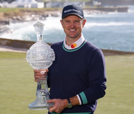 Golfer Justin Rose smiling and holding the large crystal trophy, wearing a navy blue sweater and a black cap with the Morgan Stanley logo. The ocean is visible in the background.