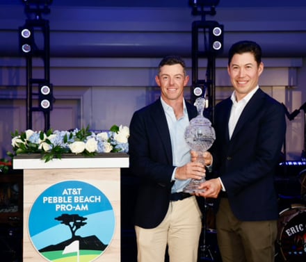 The winning Pro-Am team of Rory McIlroy and his amateur partner holding the crystal trophy on a stage, with the AT&T Pebble Beach Pro-Am logo sign nearby.