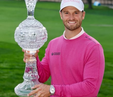 Golfer Wyndham Clark smiling and holding the large crystal trophy, wearing a bright pink sweater and a white Titleist cap.