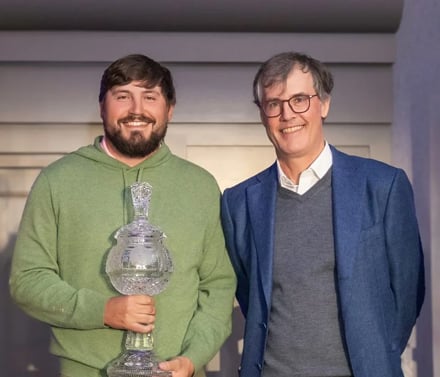 The winning Pro-Am team of a bearded amateur golfer in a green sweatshirt holding the trophy, posing next to his partner, a man in a blue blazer and glasses.