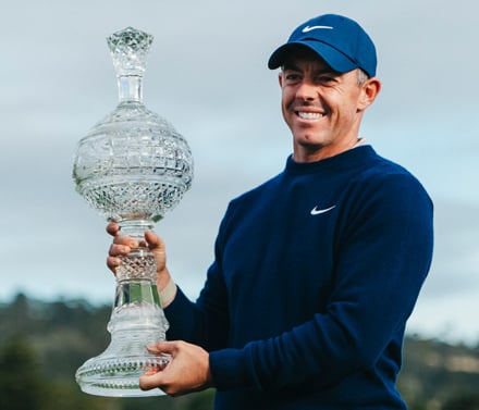 Professional golfer Rory McIlroy smiling while holding a large, ornate crystal trophy, wearing a dark blue Nike sweater and cap.