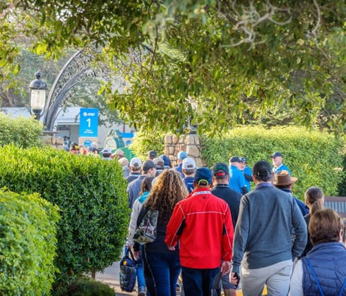 A group of spectators walking toward the Pebble Beach entrance, passing under a decorative metal archway and between well-maintained green hedges.
