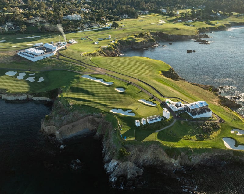A wide, high-angle aerial view of the Pebble Beach golf course on a cliff-lined peninsula, showing multiple holes, large hospitality structures, and white sand bunkers.