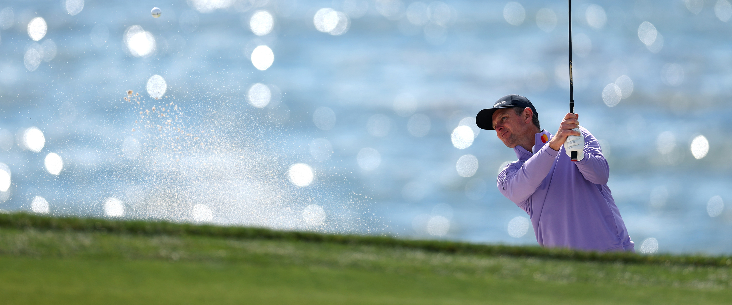 A golfer in a lavender shirt hits a shot from a sand trap, with a plume of sand rising up. The ocean water, sparkling with bright bokeh highlights, forms a soft blue background.