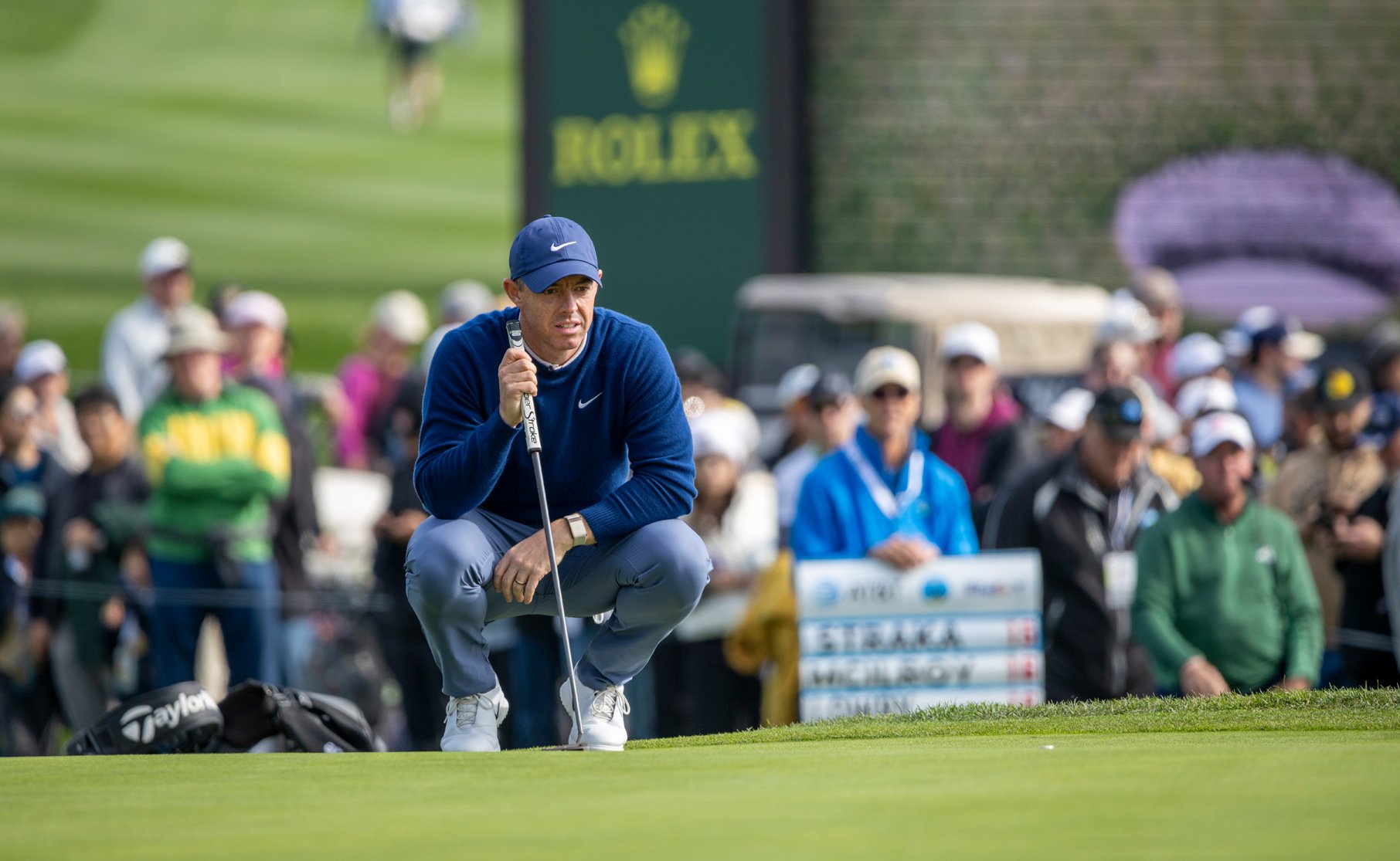 Golfer Rory McIlroy crouches low on the green to line up a putt, holding his putter. A crowd of spectators and a Rolex banner are visible in the background.