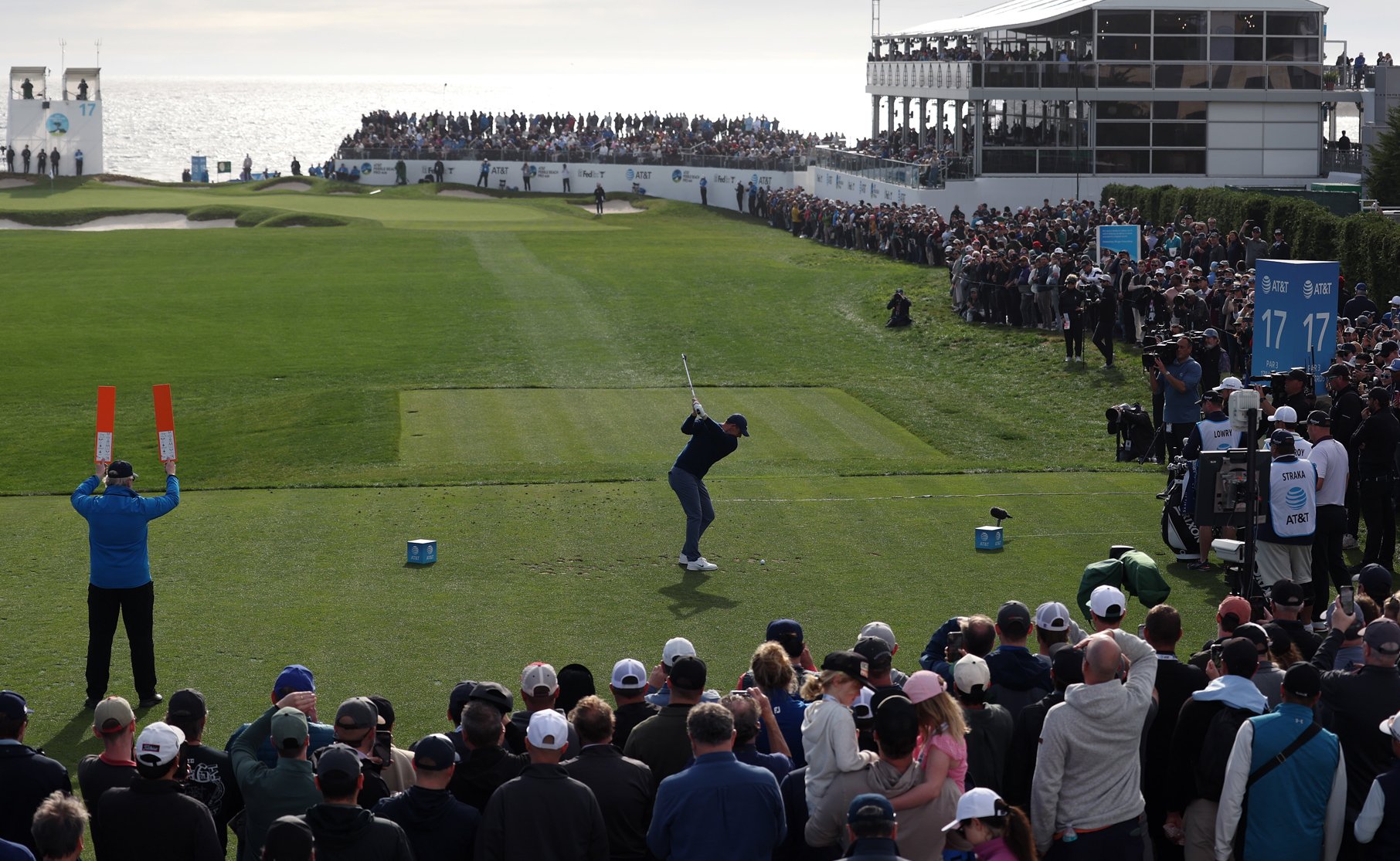 A golfer (likely Rory McIlroy) drives the ball from the tee box, surrounded by a large crowd. A marshal in the foreground holds up an orange signal board, with the 17th hole scoreboard structure and large hospitality areas visible in the distance.