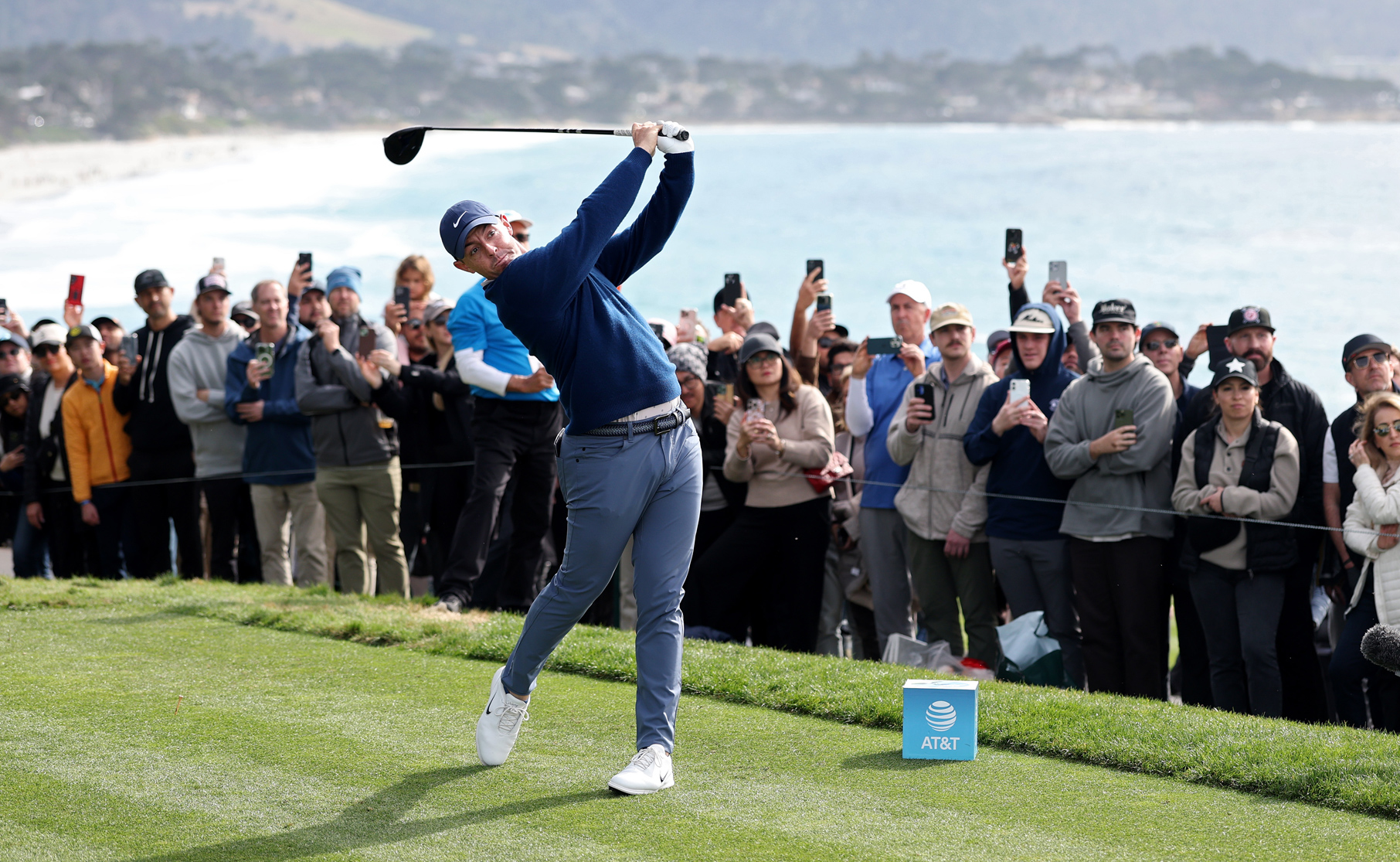 Golfer Rory McIlroy completes a powerful full-body swing from the tee box, with a large gallery of spectators behind him capturing the shot on their phones.