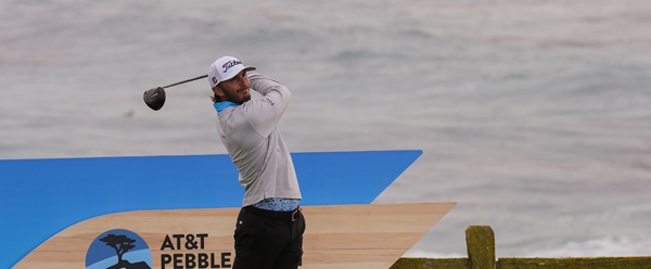 Golfer Max Homa finishes his swing after driving the ball, with the AT&T Pebble Beach Pro-Am logo sign and a wooden fence visible behind him near the overcast ocean.