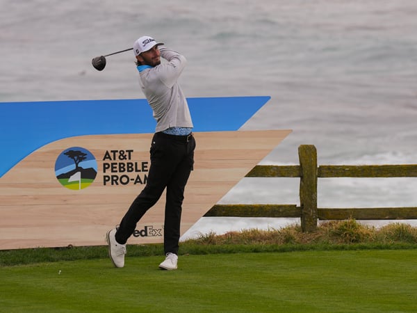 Golfer Max Homa finishes his swing after driving the ball, with the AT&T Pebble Beach Pro-Am logo sign and a wooden fence visible behind him near the overcast ocean.