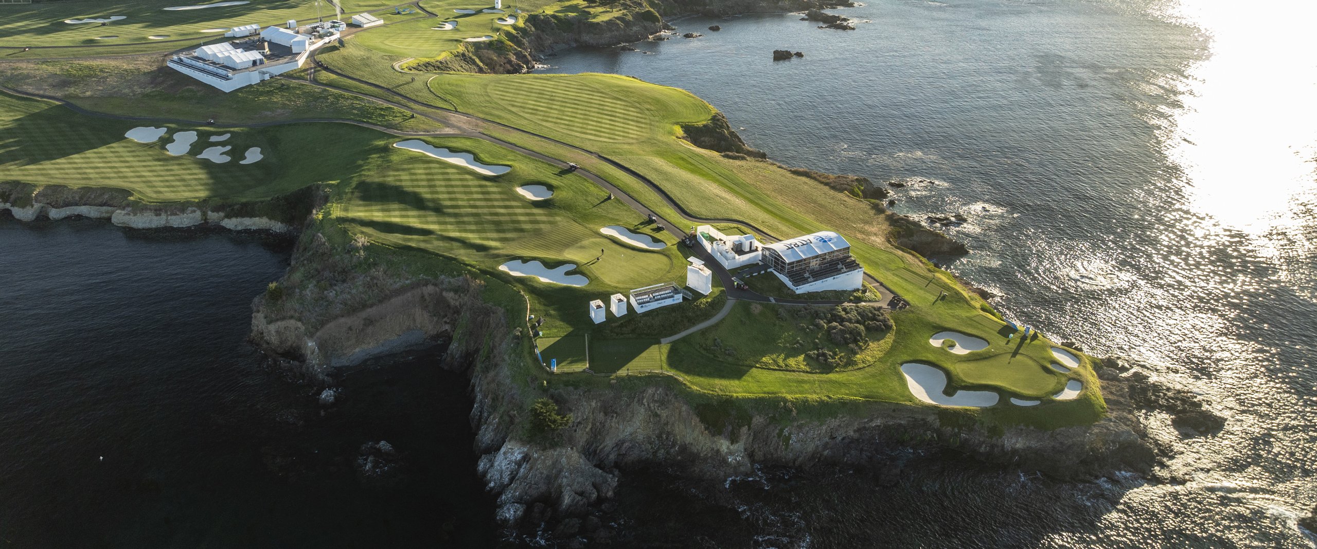 A high-angle aerial view of the Pebble Beach golf course, focusing on a rocky coastal peninsula with several holes and white hospitality structures.