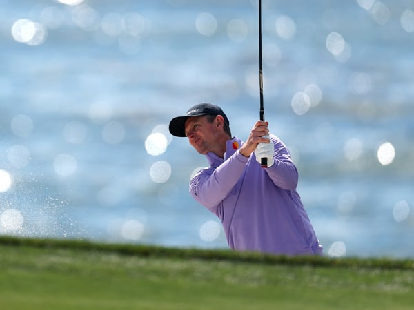 A golfer in a lavender pullover hits a shot out of a sand trap, with a plume of sand and the bright, sparkling ocean creating a bokeh effect in the background.