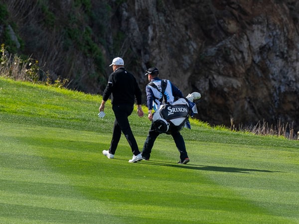 A golfer and his caddie (carrying a Srixon bag) walk across a lush green fairway, moving toward a dark, rugged cliff face in the background.