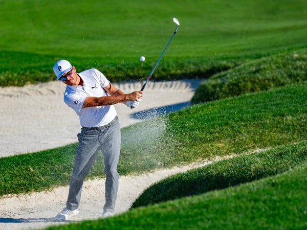 Golfer Rickie Fowler is mid-shot, hitting the ball out of a large sand bunker on a sunny day, kicking up a dramatic spray of sand.