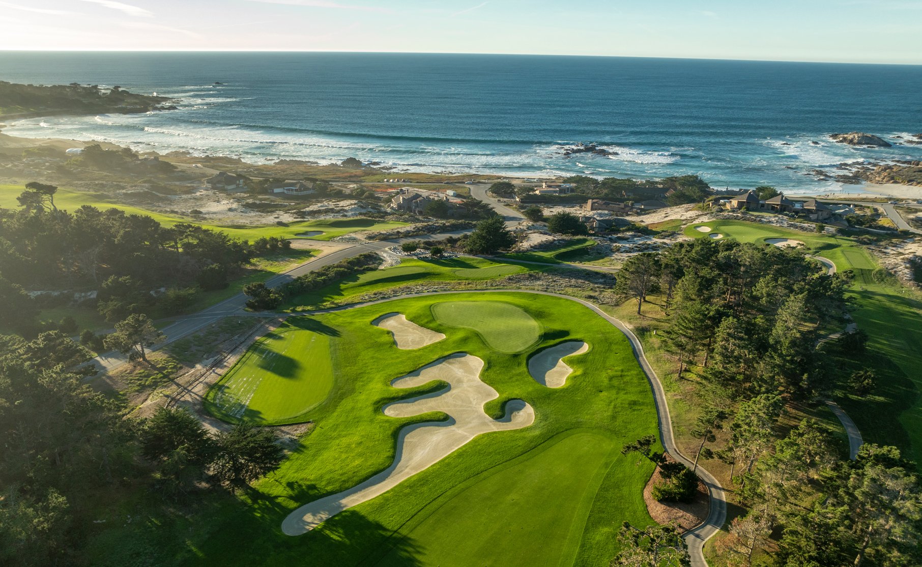 An aerial view of a golf hole at Pebble Beach with a large, intricately shaped sand trap bordering the green. Beyond the course are cypress trees, sandy coastline, and the blue ocean.