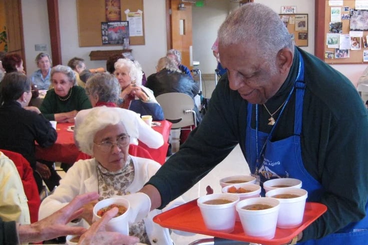 An older male volunteer in a blue apron smiles as he leans over to serve a cup of soup on a tray to an older woman seated at a table in a crowded community dining hall.