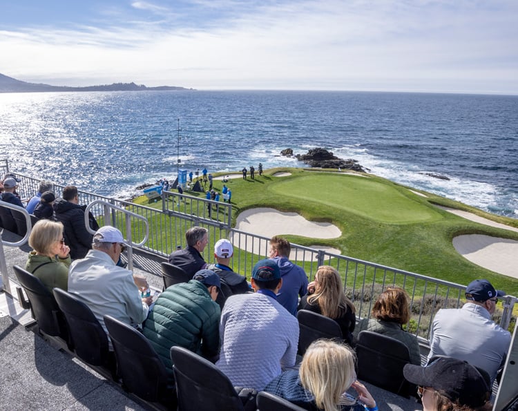 Spectators seated in tiered grandstands overlooking a cliffside golf green with large sand bunkers, set against the bright blue ocean and the far coastline.