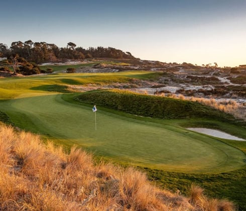 A close-up of a golf green at Spyglass Hill in the late afternoon sun, framed by golden-brown sand dunes and tall grasses.
