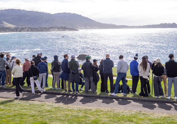 A group of young spectators (possibly students) standing along the edge of the golf course, facing the ocean, with the bright, sparkling water and coastal mountains in the background.