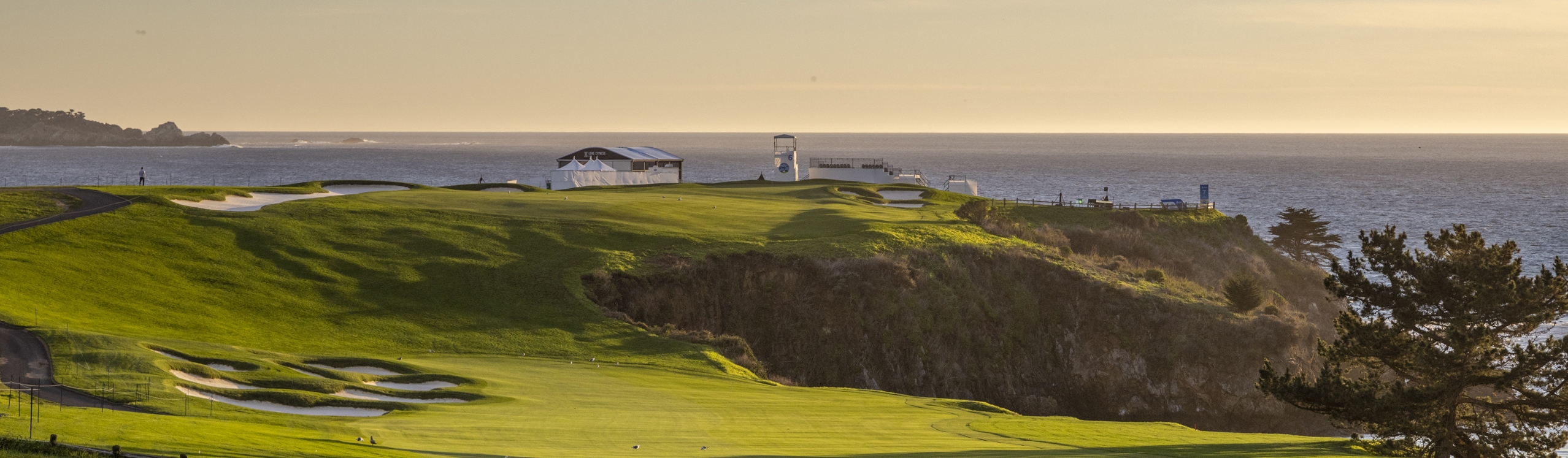 A wide view of the Pebble Beach golf course at sunset, showing the lush green fairway and rough leading up a cliffside to hospitality tents and structures overlooking the glowing ocean.
