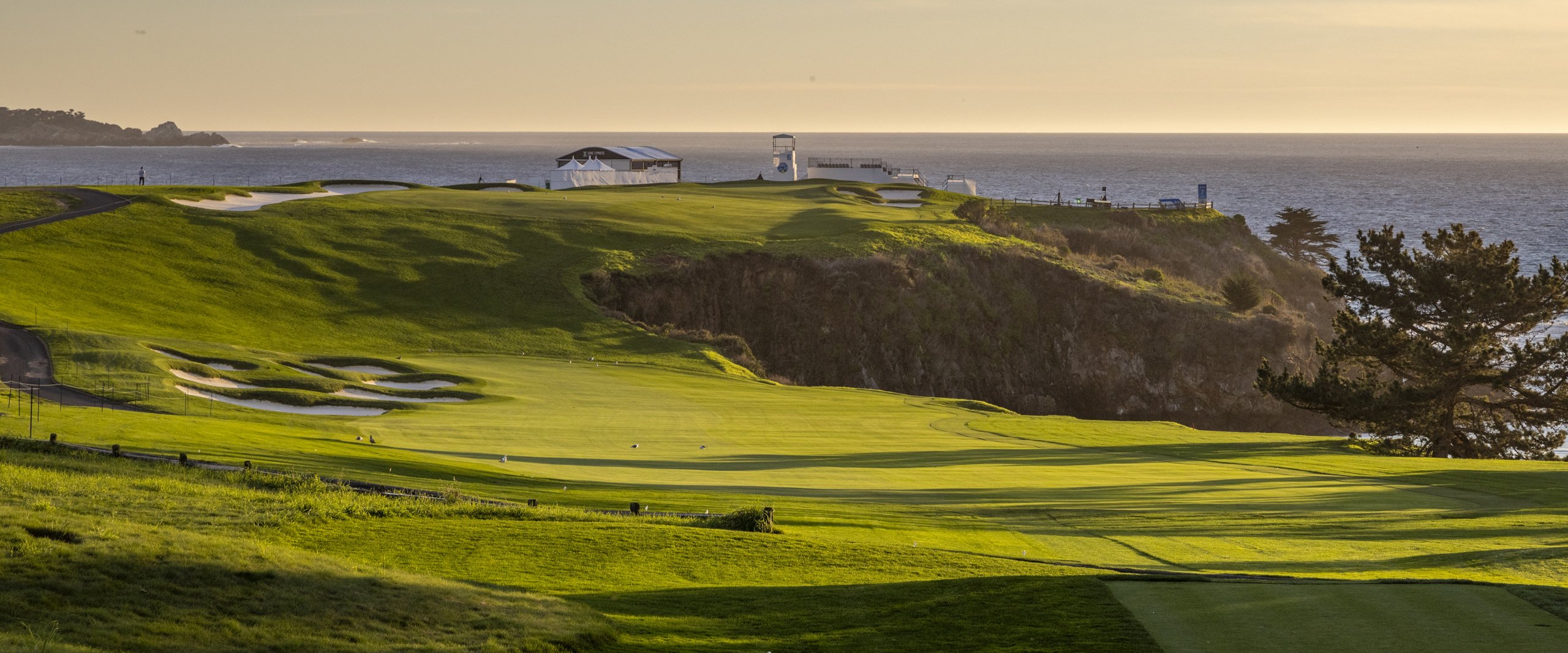 A wide view of the Pebble Beach golf course at sunset, showing the lush green fairway and rough leading up a cliffside to hospitality tents and structures overlooking the glowing ocean.