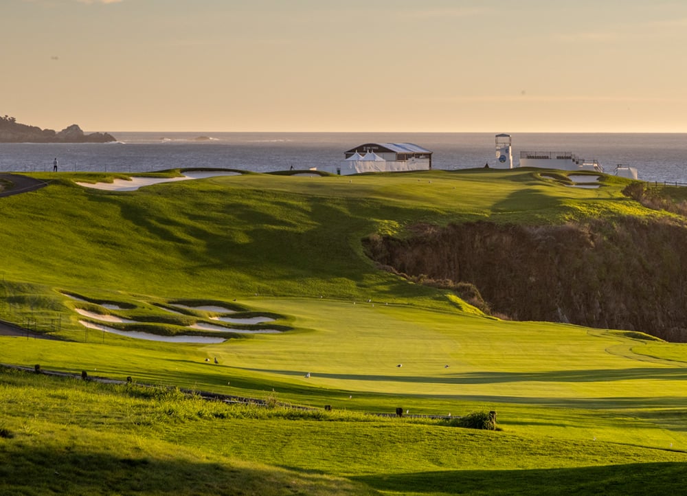 A mid-level view of a golf fairway and green at Pebble Beach at sunset, with tiered sand bunkers and large white hospitality tents overlooking the golden ocean horizon.