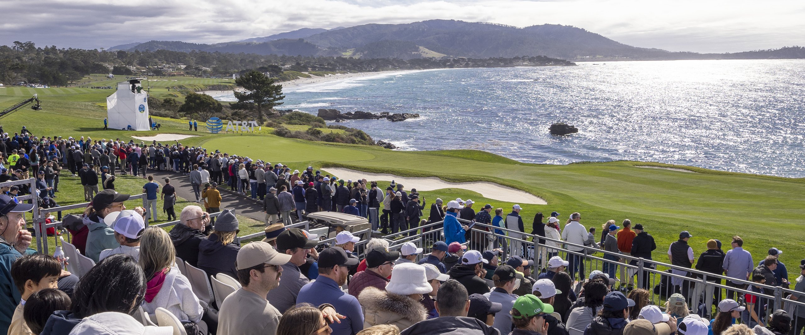 A wide view of the Pebble Beach course from a grandstand, showing a large crowd of spectators and the green fairway leading to the ocean, with mountains on the horizon.