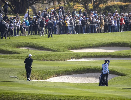 A golfer and his caddie walk past a large sand bunker with a crowd of spectators lined up on the hillside above them. The golfer is seen from behind in a black shirt.