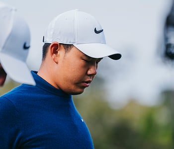 A close-up of professional golfer Tom Kim wearing a navy blue sweater and white Nike cap, looking down with a focused expression.