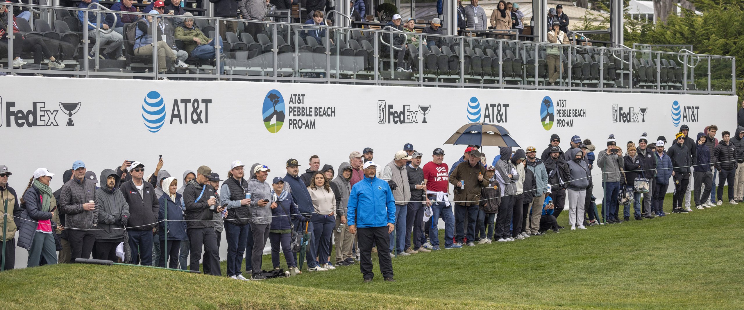 A large crowd of spectators stand in a long line along the ropes of the golf course, watching the action. A tiered grandstand with AT&T and FedEx sponsor branding is visible above them.