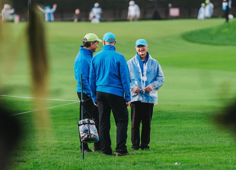 A shot of three golf volunteers in blue jackets talking together on the edge of the green, with one wearing a clear rain poncho and another a neon green cap.