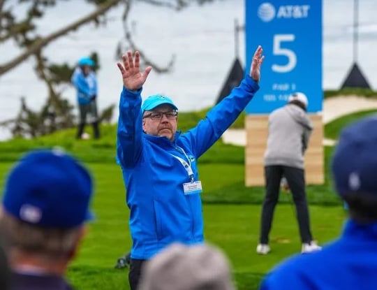 A golf course marshal (volunteer) in a bright blue jacket and cap stands on the tee box with their arms raised, signaling to the crowd. The Hole 5 scoreboard is visible behind him.