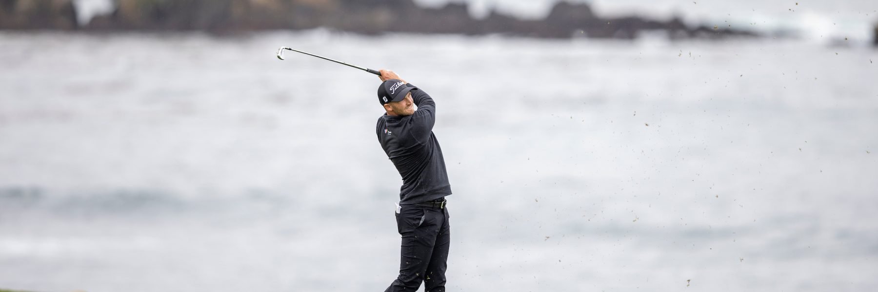 A wide shot of golfer Wyndham Clark in a black outfit, completing a powerful swing against the backdrop of a gray, overcast ocean.